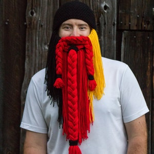 May include: A person wearing a black knit hat with a long, braided yarn beard in black, red, and yellow.