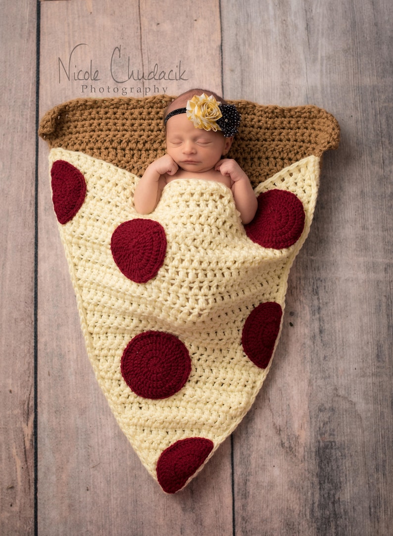 Puede incluir: Un beb&eacute; reci&eacute;n nacido est&aacute; envuelto en una manta de crochet en forma de rebanada de pizza con acentos de pepperoni rojos. El beb&eacute; lleva una diadema de flores. El texto "Nicole Chudacik Photography" es visible en la esquina superior izquierda de la imagen.
