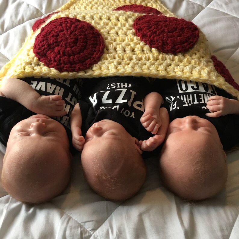 Puede incluir: Tres beb&eacute;s con monos negros con texto sobre pizza est&aacute;n tumbados bajo una manta de pizza de ganchillo. La manta es amarilla con c&iacute;rculos rojos de pepperoni.