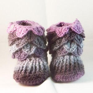 May include: A pair of crocheted baby booties in shades of purple, pink, and gray. The booties have a scalloped edge and a textured design.