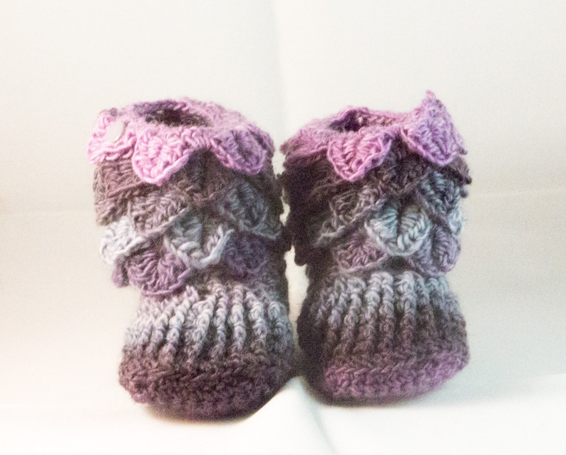 May include: A pair of crocheted baby booties in shades of purple, pink, and gray. The booties have a scalloped edge and a textured design.