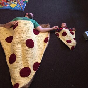 Puede incluir: Un padre y un hijo est&aacute;n acostados en mantas de crochet de rebanada de pizza a juego. Las mantas son amarillas con c&iacute;rculos de pepperoni rojos. El padre lleva una camisa verde y el hijo lleva una camisa roja.