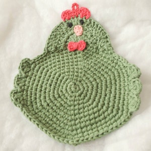 May include: Green crocheted dishcloth with a chicken face. The chicken has a pink beak, black eyes, and a red bow tie.