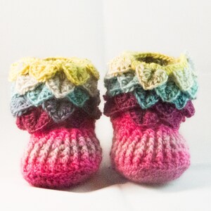 May include: A pair of crocheted baby booties with a multi-colored ruffled trim. The booties are pink with a gradient of lighter pink and white.