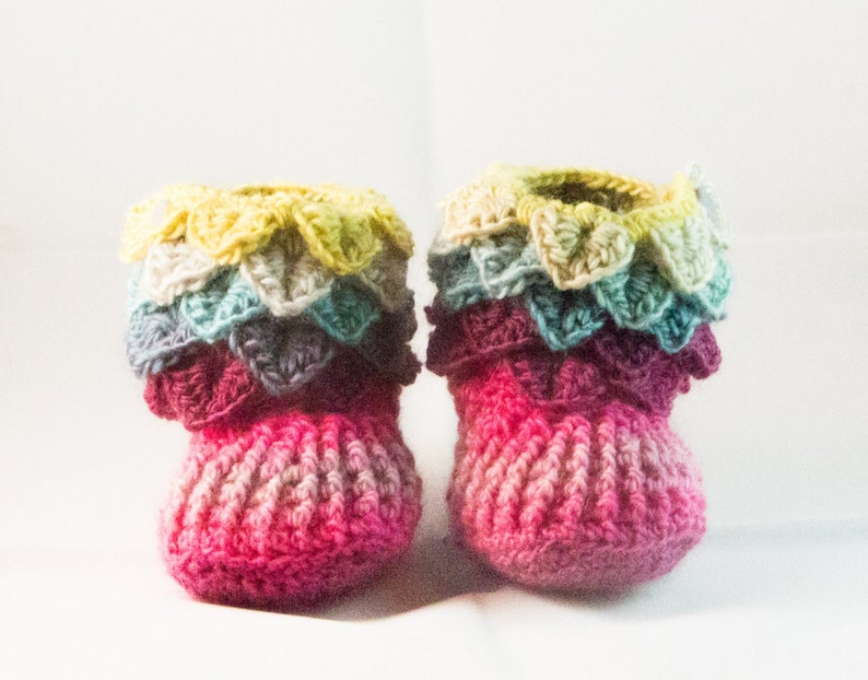 May include: A pair of crocheted baby booties with a multi-colored ruffled trim. The booties are pink with a gradient of lighter pink and white.