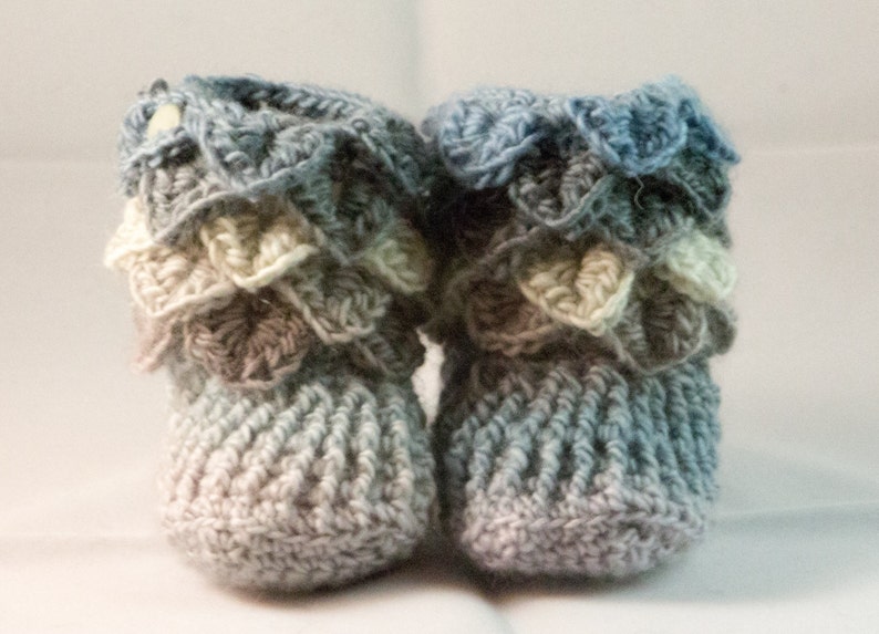 May include: A pair of crocheted baby booties in shades of blue, gray, and white. The booties have a scalloped edge and a textured knit pattern.