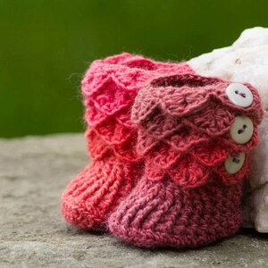 May include: A pair of pink and red crocheted baby booties with buttons. The booties are sitting on a rock.