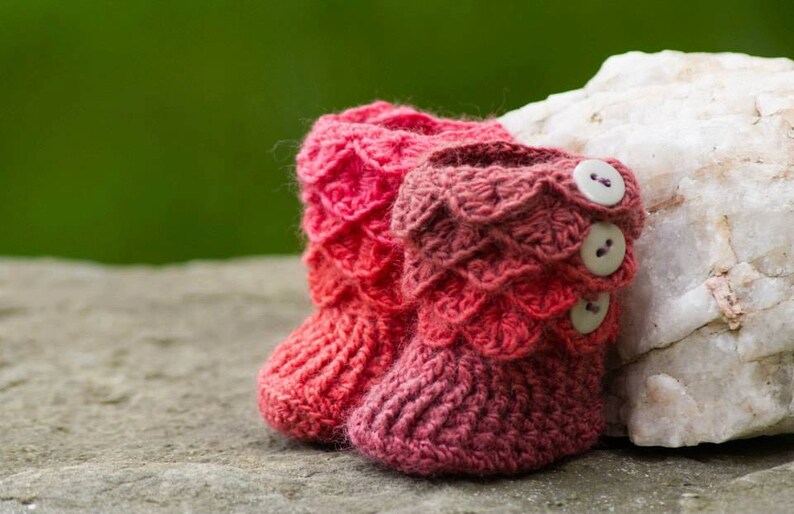 May include: A pair of pink and red crocheted baby booties with buttons. The booties are sitting on a rock.