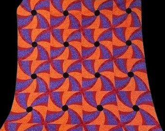 May include: A patterned fabric featuring a repeating design of orange and purple pinwheel shapes. Each pinwheel has a black center. The fabric is likely used for quilting, sewing, or other craft projects.