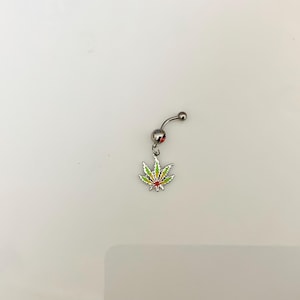 Pode incluir: Um piercing de umbigo prateado com uma barra curva e um pingente pendurado. O pingente é uma folha de cannabis colorida com detalhes em esmalte verde, amarelo e vermelho. A joia está sobre um fundo branco.
