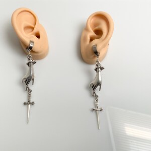 May include: A pair of silver-toned earrings. Each earring features a hand pendant holding a sword. The earrings are attached to a hoop that is clipped to a flesh-colored ear model. The background is white.