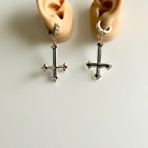 May include: Silver-toned earrings featuring an inverted cross design with red accents. The earrings are attached to a silver hoop. The earrings are displayed on a white background.