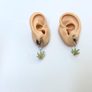 May include: A pair of silver-tone hoop earrings with dangling marijuana leaf charms. The charms feature green, yellow, and red enamel detailing. The earrings are displayed on ear models against a white background.