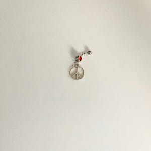 May include: A silver belly button ring featuring a peace symbol charm. The ring has a curved barbell design with a small red detail. The peace symbol is a classic design, and the ring is likely made of a durable metal.
