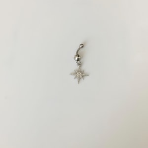 May include: A silver-colored belly button ring with a dangling starburst charm. The starburst has multiple points and is embellished with small, clear gemstones. The ring has a curved barbell design.