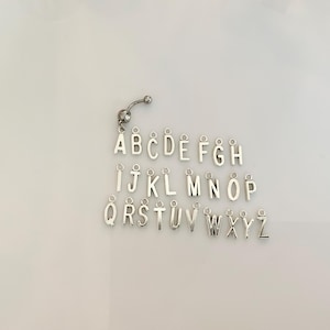 May include: A collection of silver-colored alphabet charms, each with a small loop for attachment. The letters A through Z are displayed, along with a curved barbell. The charms are arranged on a white surface.