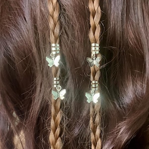 May include: Close-up of two brown braided hair strands adorned with silver butterfly charms. The braids are decorated with silver metal rings. The background is a darker shade of brown hair.
