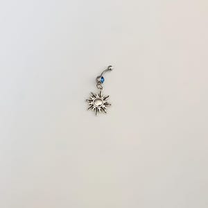 Könnte beinhalten: Ein silberner Bauchnabelpiercing mit einem Sonnen-Anhänger. Der Anhänger hat ein detailliertes Sonnendesign mit spitzen Strahlen. Über der Sonne ist ein kleiner blauer Edelstein gesetzt. Der Schmuck wird vor weißem Hintergrund präsentiert.