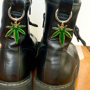 May include: Two black leather boots with chunky soles. Each boot has a gold-coloured charm shaped like a green cannabis leaf. The charms are attached to the boots with a silver-coloured ring.