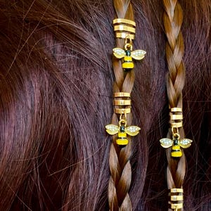 May include: Close-up of plaited brown hair decorated with gold hair cuffs and bee charms. The bee charms are yellow and black with rhinestone wings, adding a decorative element to the hairstyle.