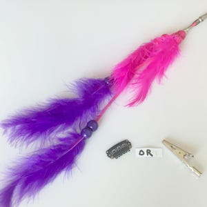 Peut inclure: Un accessoire pour cheveux avec des plumes violettes et roses, orné de perles violettes. Il comprend une pince métallique et une petite pince à cheveux noire. Le mot "OR" est écrit sur un petit rectangle blanc.