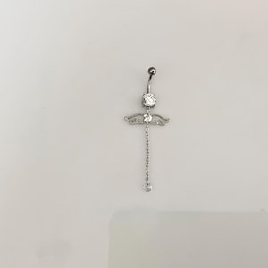 Könnte beinhalten: Ein silberfarbener Bauchnabelpiercing mit einer herabhängenden Kette und einem kleinen, klaren Edelstein. Der Ring hat ein dekoratives Design mit kleinen Flügeln und einem großen, klaren Edelstein oben.