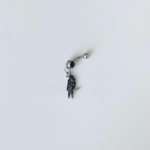 May include: A silver-toned curved barbell belly ring featuring a small dark blue circular gem on the bottom ball and a dangling charm shaped like a black bird with white details. The jewelry piece has a smooth metallic finish and a classic design.