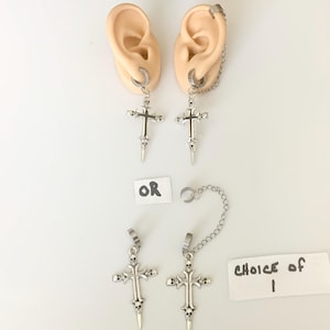 Può includere: Orecchini a croce color argento con dettagli a teschio e fondo appuntito. Un paio è mostrato attaccato a un orecchino a cerchio e un altro paio è mostrato con una catena e un ear cuff. Il testo "OR" e "CHOICE of 1" sono visibili.