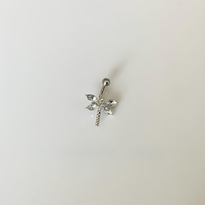 May include: A silver-toned belly button ring featuring a dragonfly design. The dragonfly has four clear, marquise-cut crystal wings and a segmented tail. A small, round clear crystal is set at the head, and a smooth metal ball sits at the top.