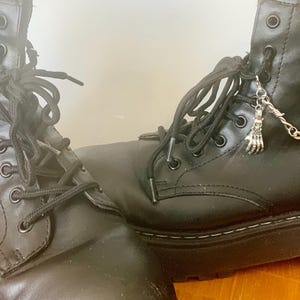 May include: Close-up of two black combat boots with black laces. Each boot has a silver chain with a small hand charm and barbed wire accents. The boots are on a wooden surface.