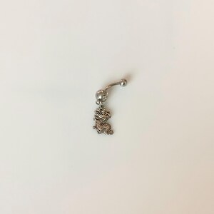 May include: A silver-toned curved barbell belly button ring featuring a small, detailed dragon charm dangling from the bottom ball. The jewelry piece includes a top ball and a larger bottom ball with a clear stone setting above the dragon charm.