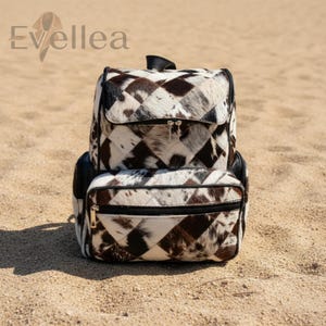 May include: A cowhide backpack with a diamond pattern in shades of brown and white. The backpack has a zippered front pocket and a top handle. The brand name "Evellea" is visible in the upper left corner.