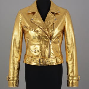 Peut inclure: Veste en cuir dorée métallisée de style motard classique. Elle présente une fermeture éclair asymétrique, un col cranté, une petite poche à monnaie et une ceinture à la taille. La veste a des manches longues avec des poignets zippés.