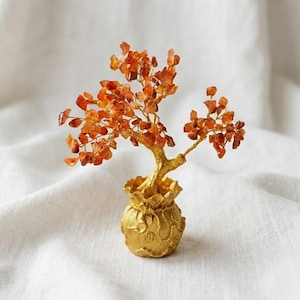 Puede incluir: Una escultura de árbol decorativa con numerosas pequeñas piedras preciosas de color naranja translúcido unidas a finas ramas de alambre dorado. El árbol está anclado en una base dorada texturizada con forma de pequeña bolsa.