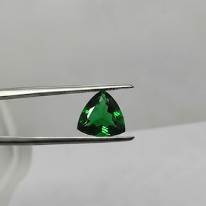 Könnte beinhalten: Ein leuchtend grüner, dreieckig geschliffener Edelstein wird von einer silbernen Pinzette gehalten. Die Facetten des Steins reflektieren das Licht und zeigen seine Klarheit und Brillanz. Der Hintergrund ist ein weiches Weiß, das die Farbe und Form des Steins hervorhebt.