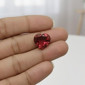 Puede incluir: Una gema facetada en forma de corazón, de un tono rojo intenso. La gema se sostiene en una mano, mostrando sus superficies pulidas y cortes intrincados. El fondo está borroso, enfatizando el color y la forma de la gema.