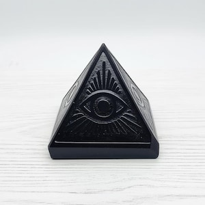 May include: A black pyramid-shaped decorative object with an eye symbol on the front. The pyramid has a flat base and is made of a solid, dark material. The eye symbol is detailed with radiating lines.