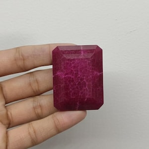 Könnte beinhalten: Ein facettierter, rechteckiger Edelstein in tiefem Magenta. Die Oberfläche des Steins weist ein Netzwerk feiner, rissartiger Einschlüsse auf, die ihm eine einzigartige Textur verleihen. Der Edelstein wird in einer Hand vor einem schlichten weißen Hintergrund gehalten.