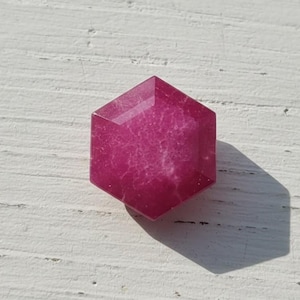 Könnte beinhalten: Ein facettierter, sechseckiger Edelstein in tiefem Magenta. Der Stein liegt auf einer weißen, strukturierten Oberfläche, mit einem Schatten nach rechts. Der Schliff des Edelsteins zeigt mehrere Facetten.