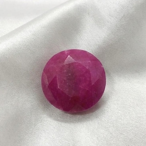 May include: A faceted, round, deep pink gemstone. The gem has multiple facets that catch the light, creating a sparkling effect. The stone is set against a white, silky background.
