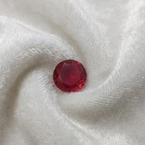 May include: A round, faceted, deep red gemstone is displayed on a soft, white, textured fabric. The gemstone's cut allows light to reflect, creating a brilliant appearance. The gem is a vibrant focal point against the neutral background.