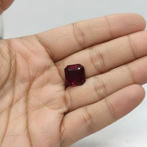 May include: A faceted, rectangular-cut, deep red gemstone. The gem is held in a hand, showcasing its rich color and clarity. The stone's facets reflect light, highlighting its brilliance. The gem appears to be a ruby, suitable for jewelry.