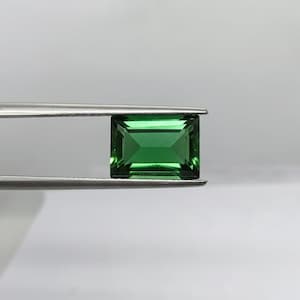 Op de afbeelding: Een rechthoekige, smaragdgroene edelsteen, vastgehouden door een zilveren pincet. De edelsteen heeft een facetgeslepen slijpsel, dat licht reflecteert. De achtergrond is een zacht, neutraal wit, dat de levendige kleur van de steen benadrukt. De edelsteen is waarschijnlijk voor sieraden.