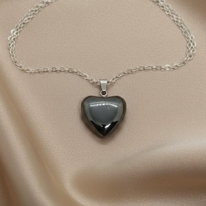 May include: A silver chain necklace with a heart-shaped pendant. The pendant is a dark, glossy gray color. The necklace is displayed on a beige satin fabric.