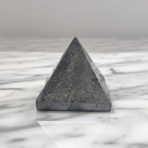 May include: A dark gray pyramid-shaped decorative object. The pyramid has a textured surface and is resting on a reflective surface. The pyramid is a geometric shape and could be used as a paperweight or decorative item.
