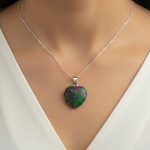 May include: A silver chain necklace with a heart-shaped pendant. The pendant is a mix of green and red colours. The necklace is worn against a white background.