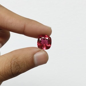 May include: A cushion-cut, faceted gemstone in a vibrant red hue. The gem is held between two fingers against a plain white background, showcasing its clarity and color. The gemstone's facets reflect light, creating a sparkling effect.