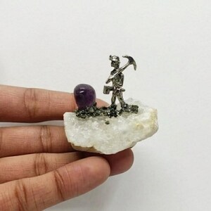 May include: A decorative figurine of a silver miner with a pickaxe, on a white quartz base. A purple amethyst gemstone and pyrite accents are also present. The figurine is held in a hand against a white background.