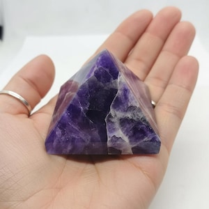May include: A pyramid-shaped amethyst crystal, showcasing a deep purple hue with white veining. The polished gemstone is held in an open hand, highlighting its geometric form and natural patterns. This crystal is a decorative item.