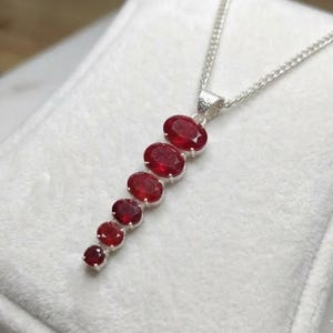 May include: A silver necklace featuring a vertical pendant composed of six graduated oval-cut red gemstones arranged in a descending line. The chain is a thin silver link style, and the stones are set in silver metal settings with a textured bail.